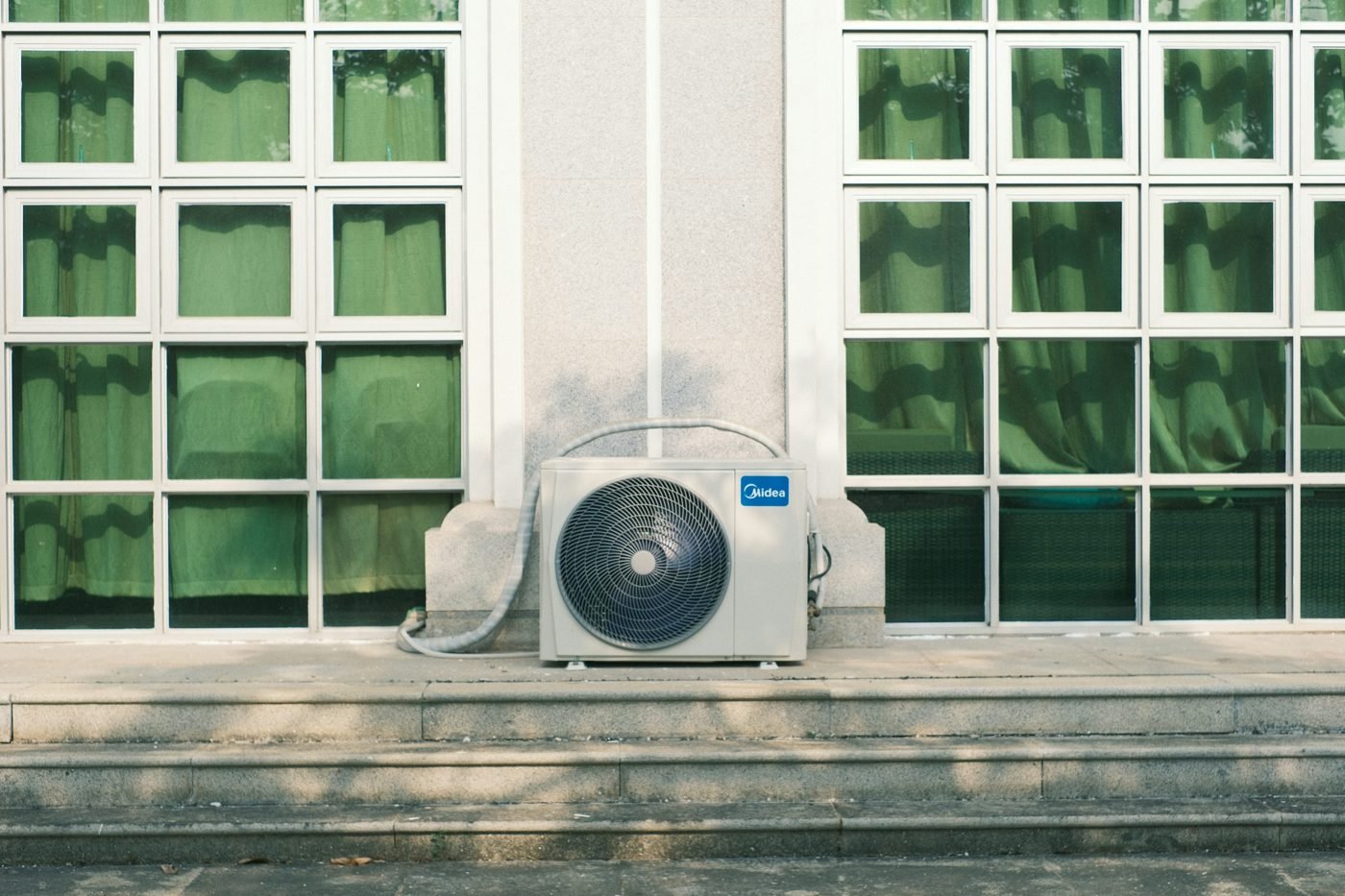 Outdoor air conditioning unit below two windows with green curtains.