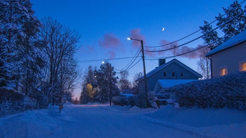 Snow-covered residential street at dusk beneath a crescent moon.
