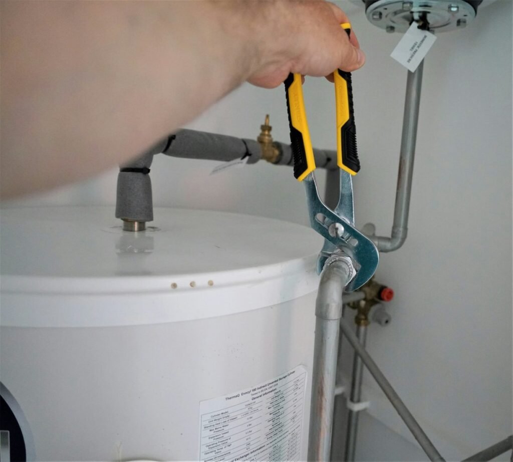 Hand using plumbing pliers on a pipe connection above a water heater.