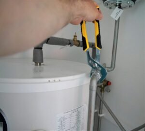 Hand using plumbing pliers on a pipe connection above a water heater.